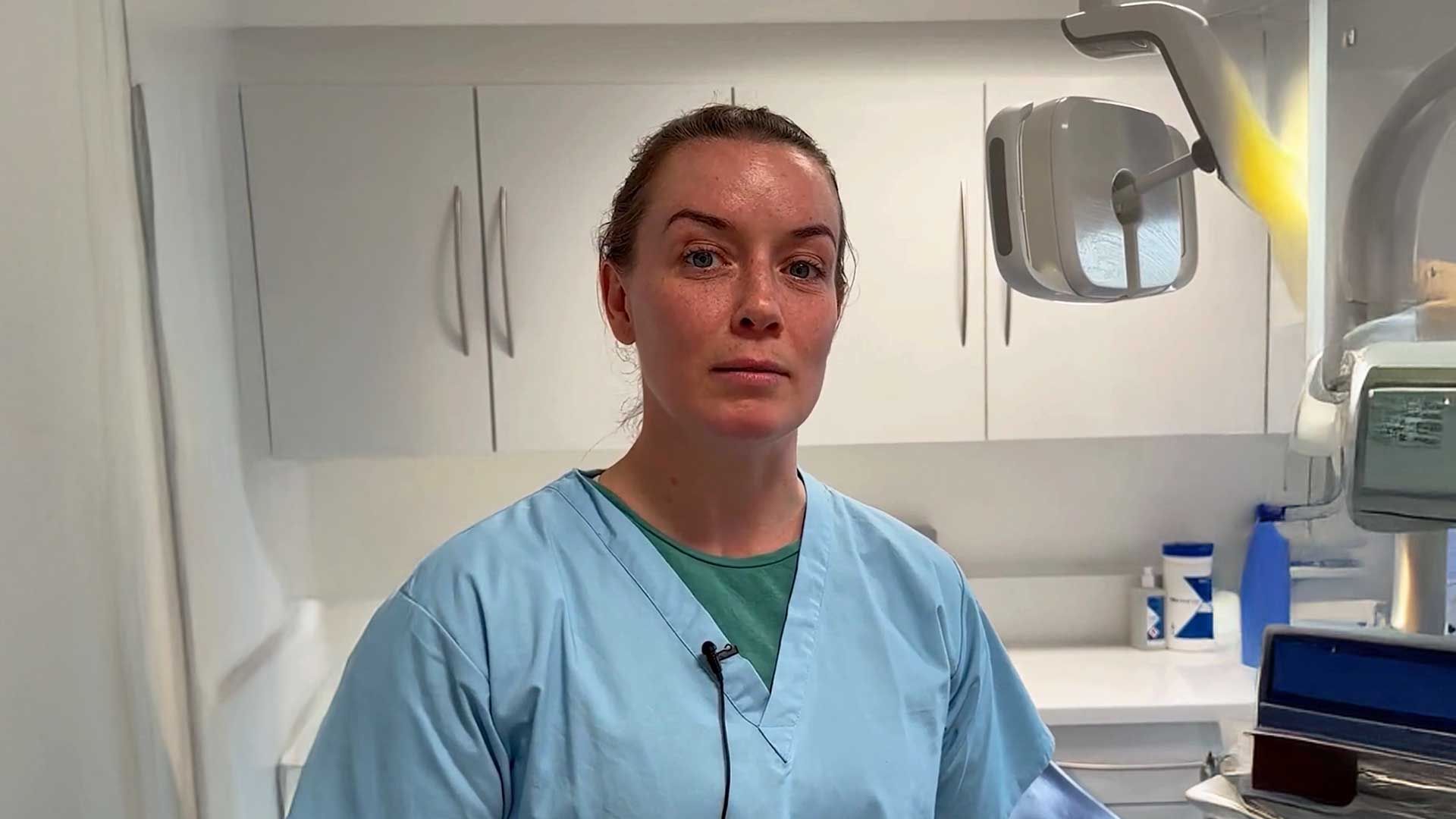 A woman in blue scrubs stands in a dental office, with cabinets and dental equipment in the background.