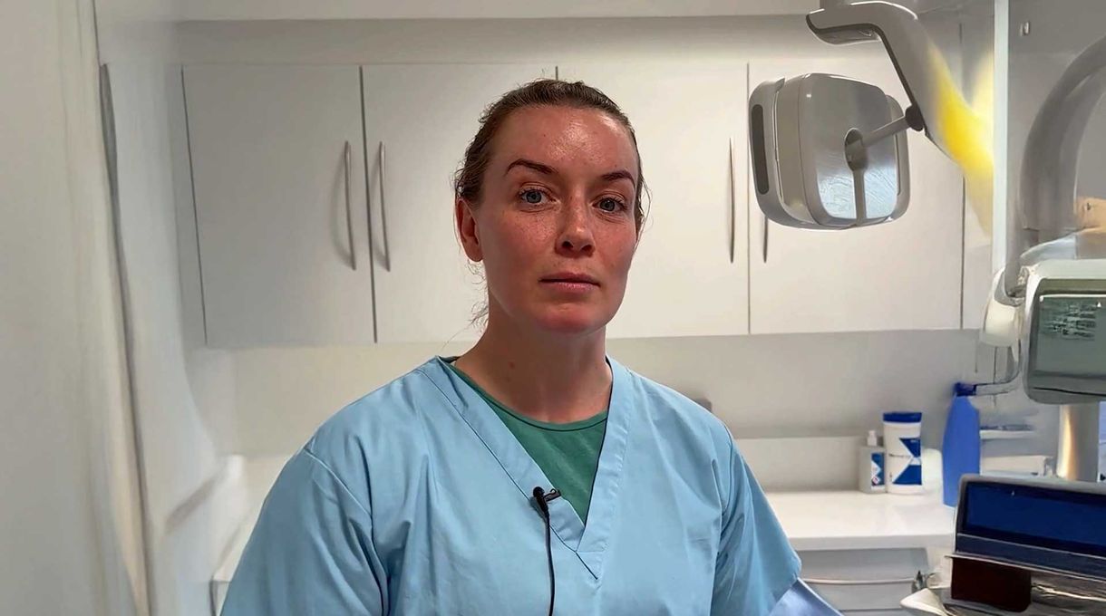 A woman in blue scrubs stands in a dental office, with cabinets and dental equipment in the background.