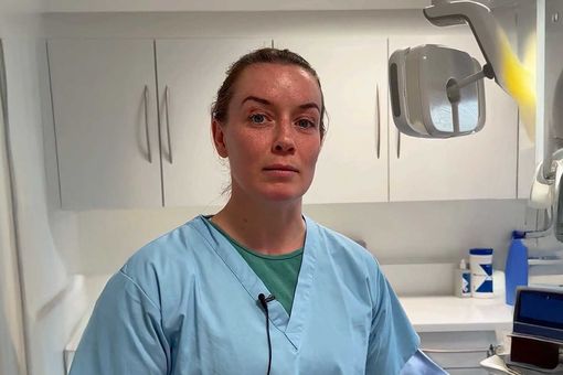 A woman in blue scrubs stands in a dental office, with cabinets and dental equipment in the background.