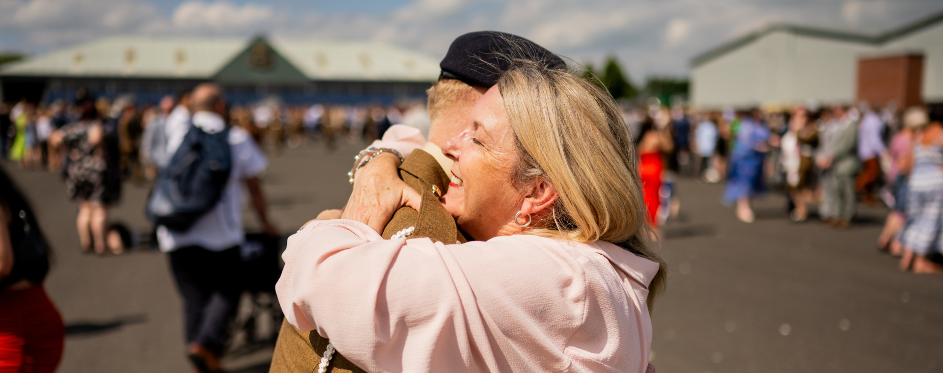 Two people embrace warmly in an outdoor setting, with a crowd and buildings blurred in the background under a partly cloudy sky.