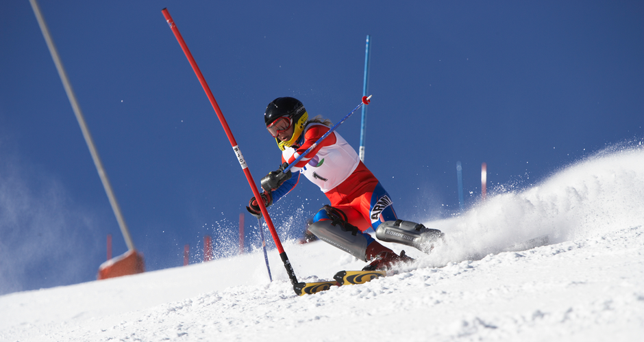 A skier in action during a slalom race on a snowy slope, with poles marking the course and snow flying around, under a clear blue sky.