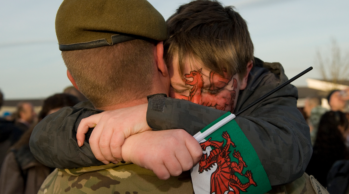 Child with dragon face paint hugs a soldier, holding a Welsh flag, during an emotional reunion outdoors.
