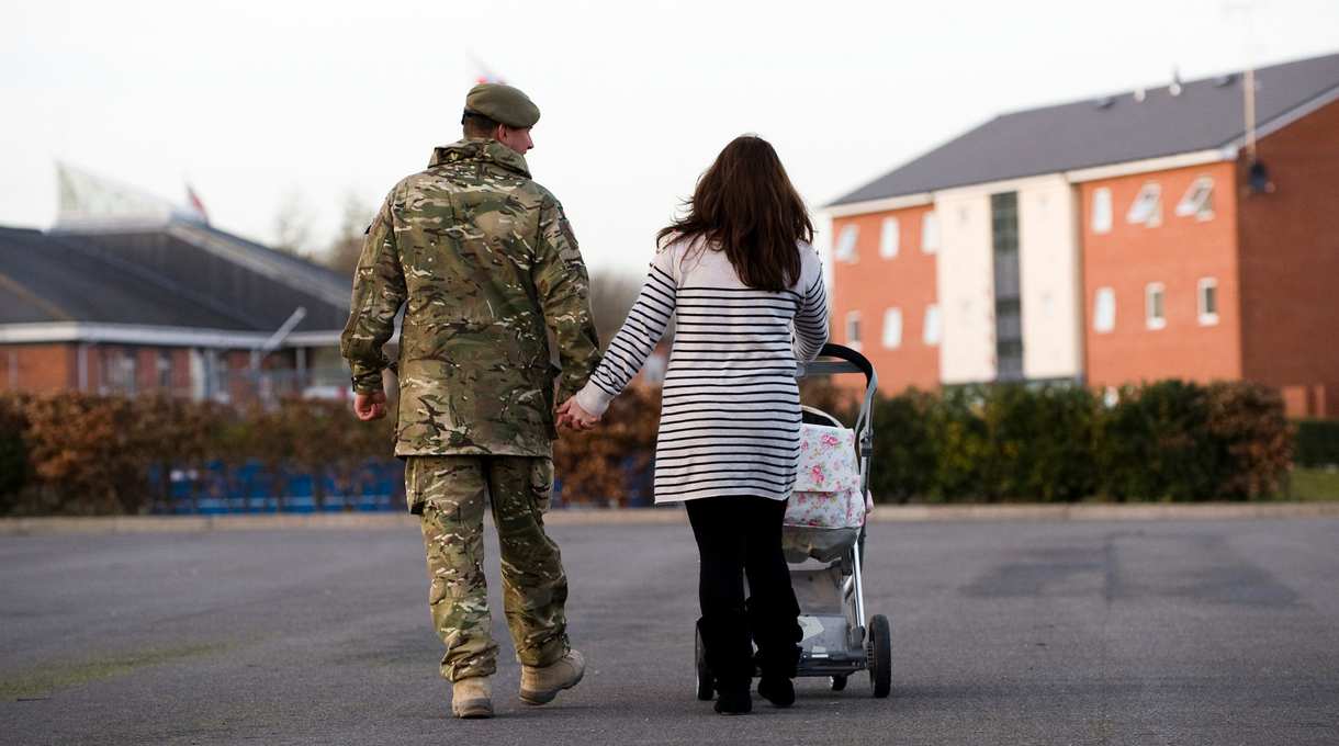 A soldier in uniform walks hand in hand with a woman pushing a stroller on a pathway near residential buildings.