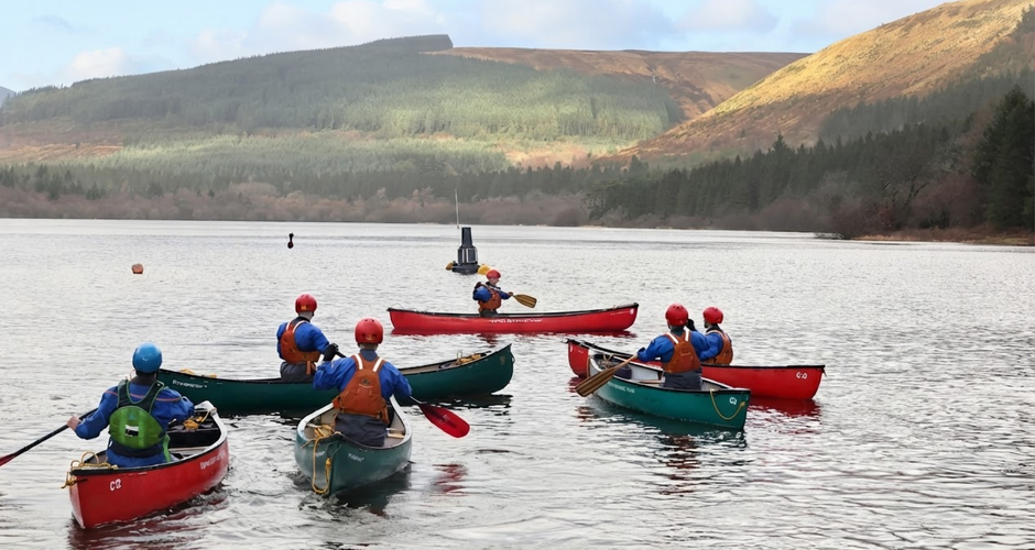 A group of people in colorful canoes paddling on a lake surrounded by scenic hills and forests under a partly cloudy sky.