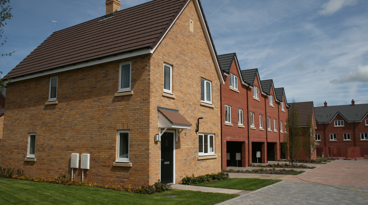 A row of modern brick houses with sloped roofs and paved driveways under a blue sky with clouds.