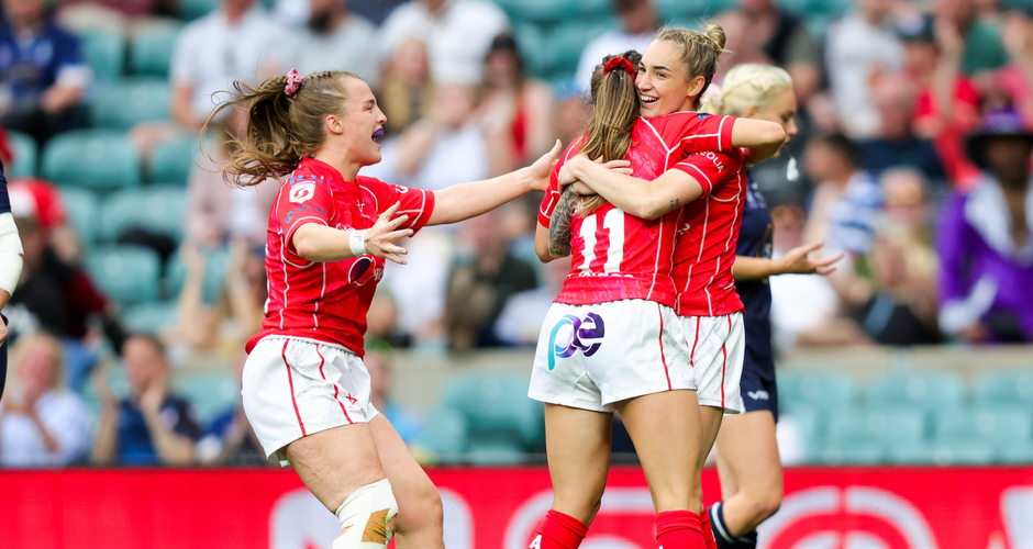 Four female rugby players in red jerseys celebrate together on the field, embracing joyfully in a stadium with an audience in the background.