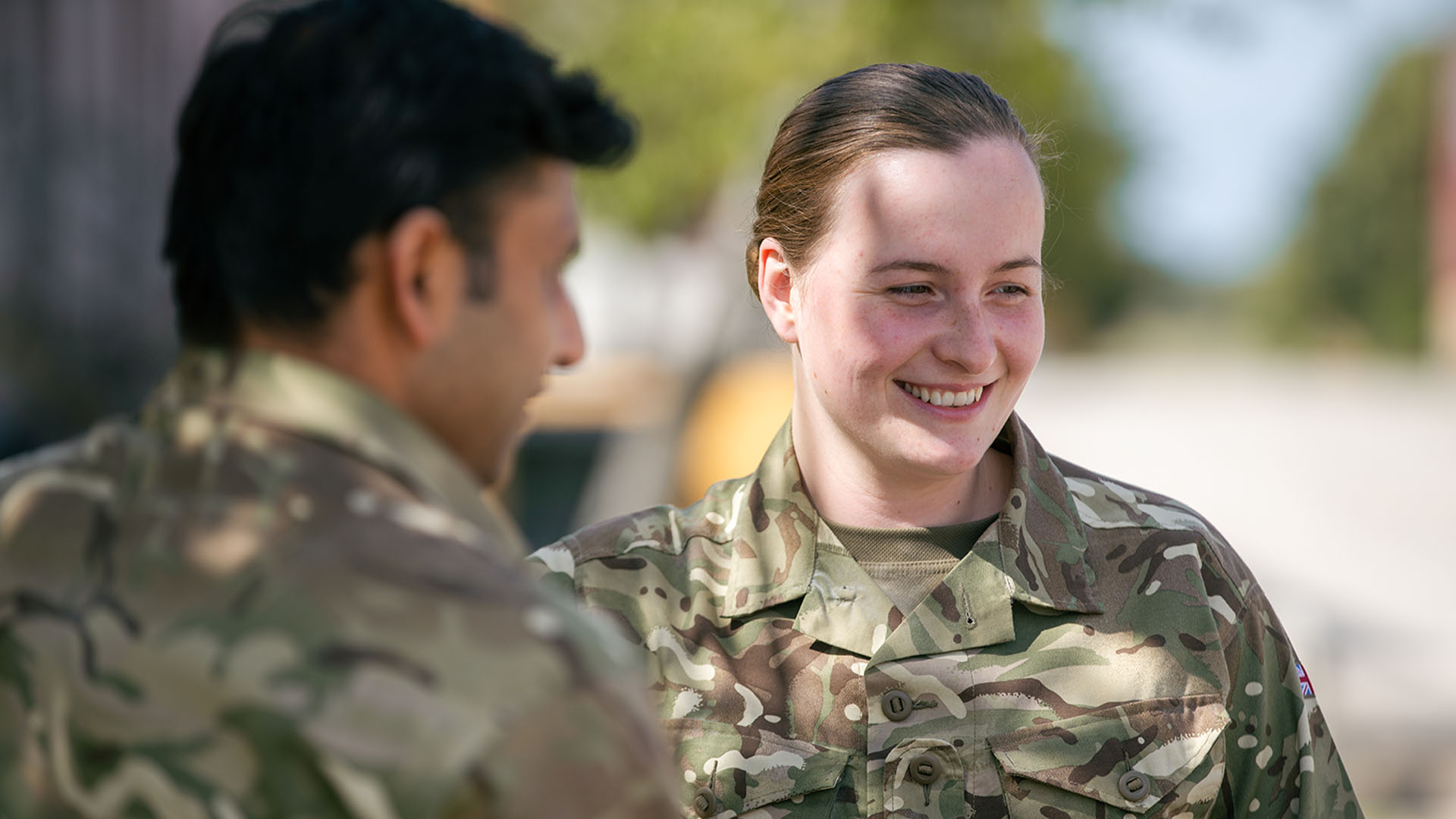 Two people in camouflage uniforms conversing outdoors, with one smiling. Trees and a clear sky in the background.