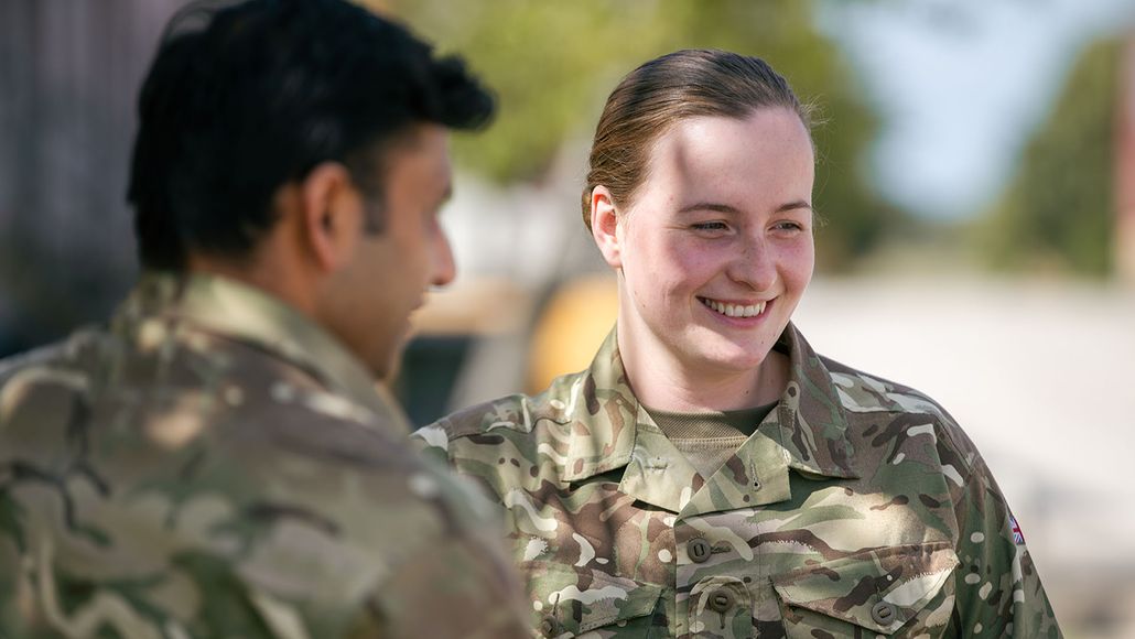 Two people in camouflage uniforms conversing outdoors, with one smiling. Trees and a clear sky in the background.