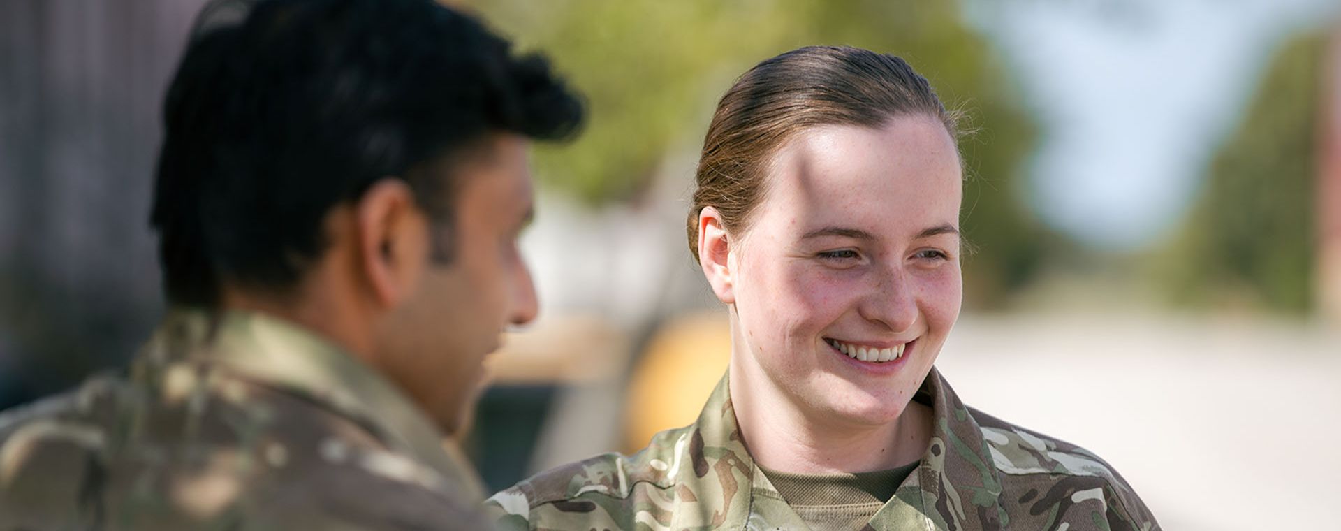 Two people in camouflage uniforms conversing outdoors, with one smiling. Trees and a clear sky in the background.