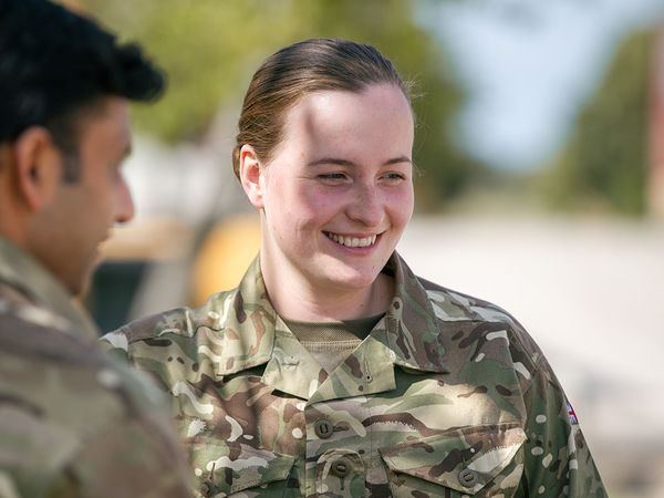 Two people in camouflage uniforms conversing outdoors, with one smiling. Trees and a clear sky in the background.