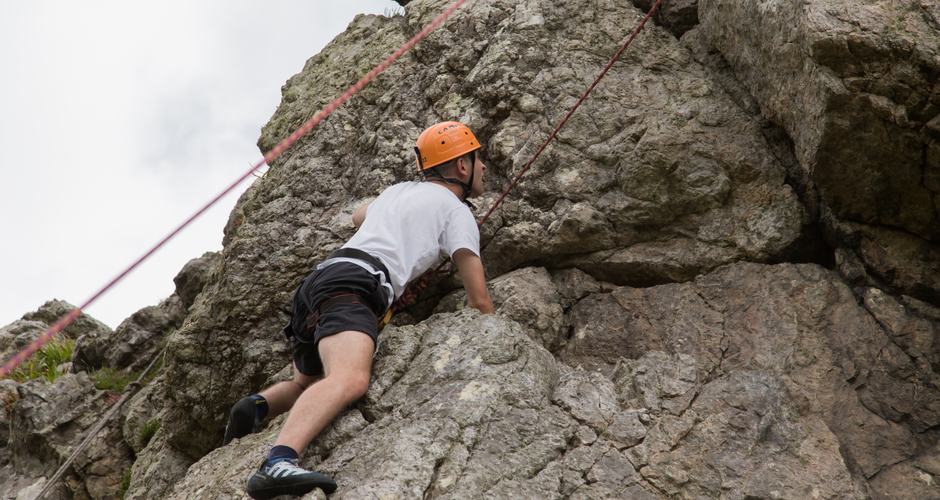 A person wearing a helmet and climbing gear scales a rocky cliff, secured by a red rope against a cloudy sky.