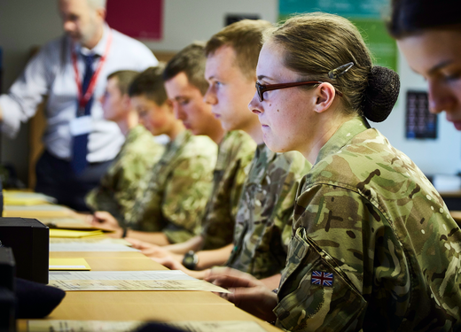 A group of soldiers in camouflage uniforms sit attentively at desks, taking notes in a classroom setting.