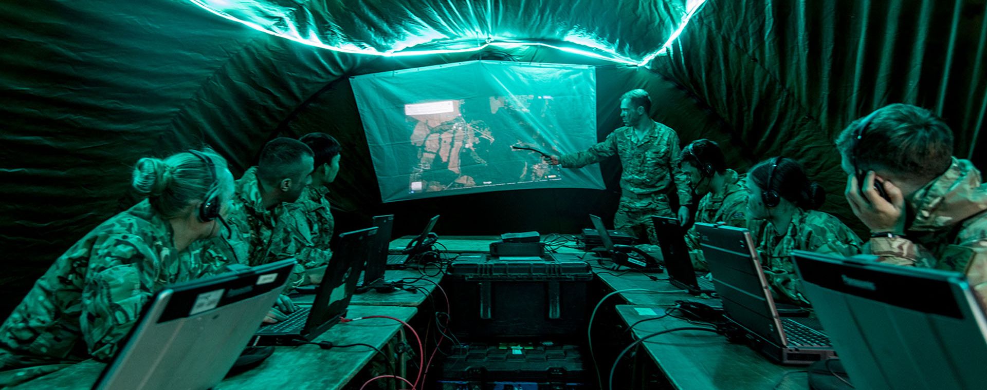 Soldiers in camouflage uniforms inside a dimly lit tent, observing a presentation projected on a screen.