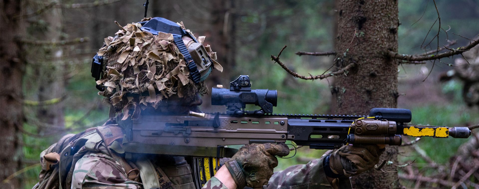 A soldier in camouflage gear and a helmet aims a rifle in a forested area, surrounded by trees and greenery.