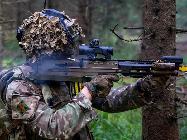 A soldier in camouflage gear and a helmet aims a rifle in a forested area, surrounded by trees and greenery.