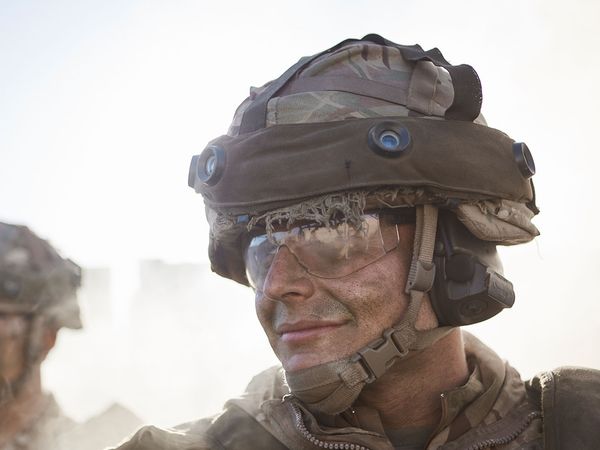Soldier in camouflage gear and helmet with goggles, smiling amidst a dusty background, another soldier slightly visible in the background.