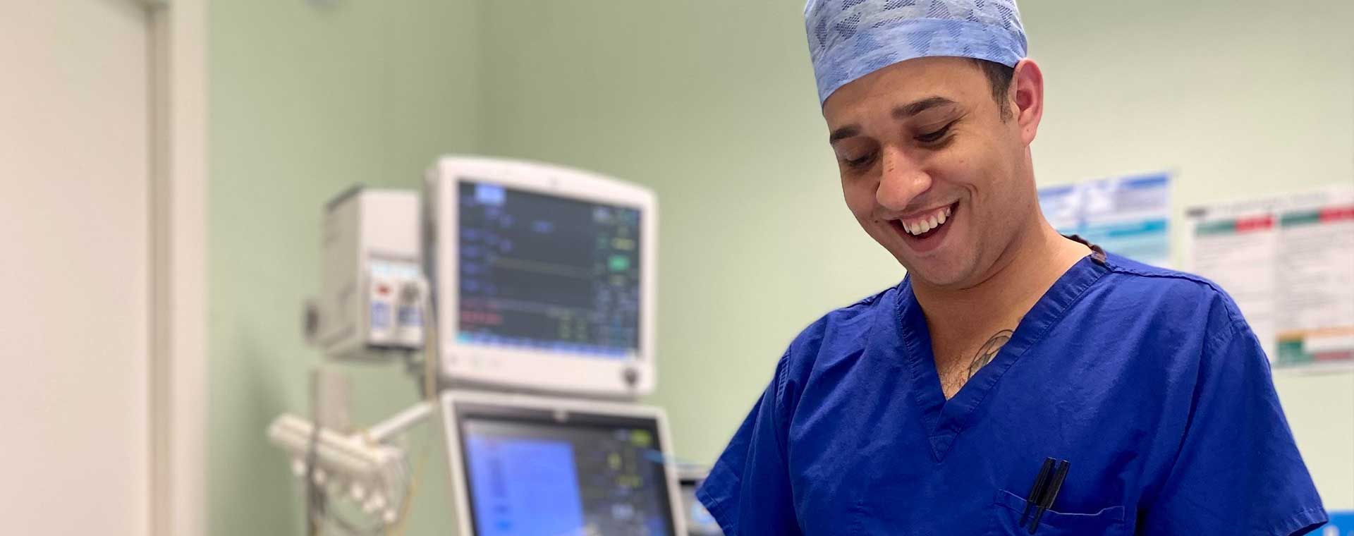 Smiling medical professional in blue scrubs and gloves prepares equipment in a hospital room with monitors in the background.