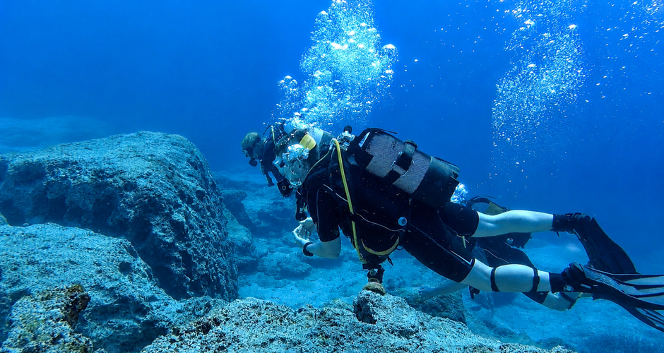 Two scuba divers exploring underwater rocks, with air bubbles rising towards the surface in clear blue water.