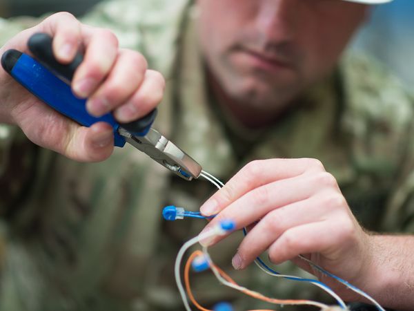 Close-up of a person in camouflage holding pliers, working on blue wires, focused on precise handling.