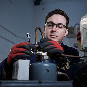 Technician wearing safety glasses and gloves works on industrial equipment in a workshop, adjusting a valve on machinery.