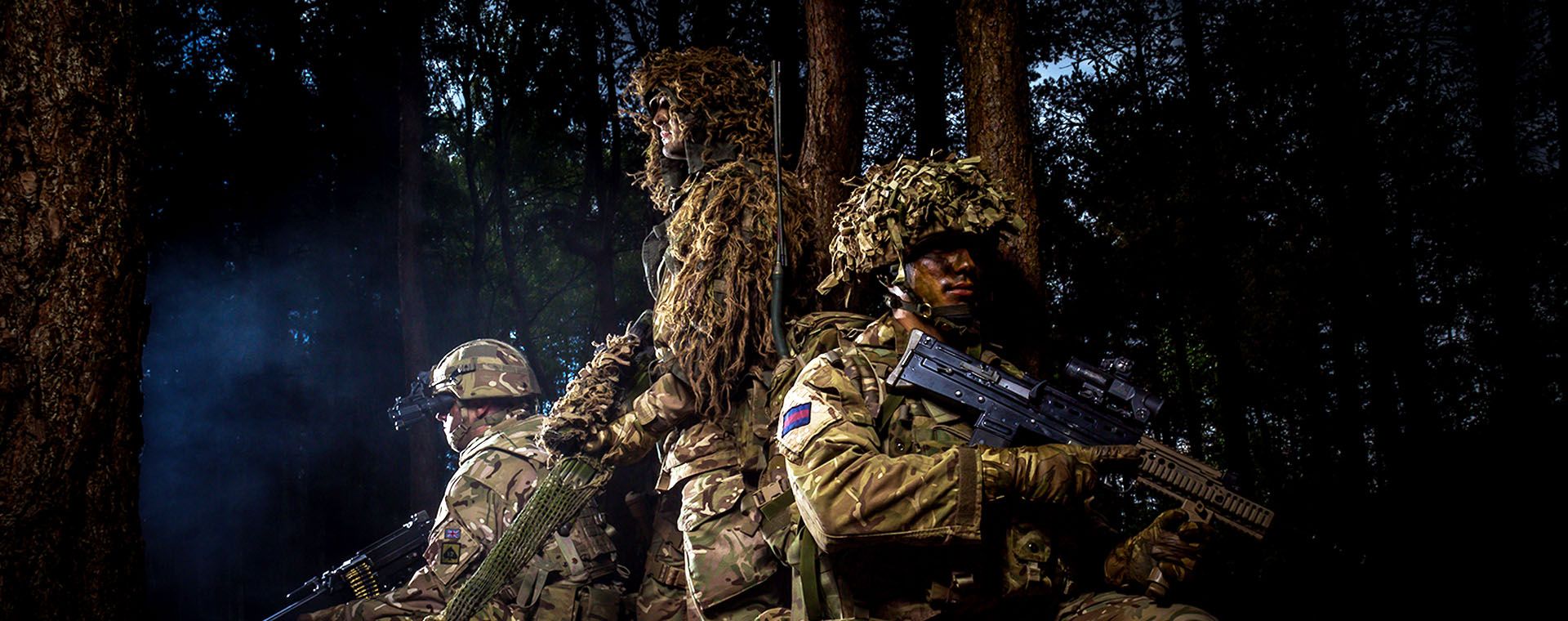 Three soldiers in camouflage and gear holding rifles, standing and kneeling in a dark forest setting.