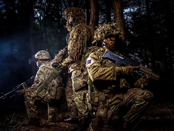 Three soldiers in camouflage and gear holding rifles, standing and kneeling in a dark forest setting.