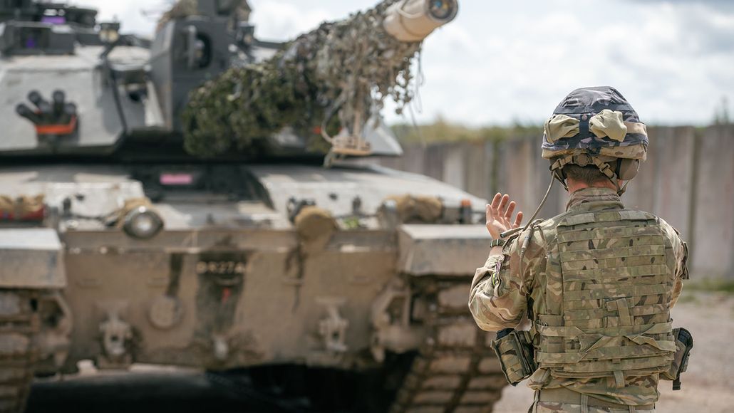 A soldier in camouflage uniform signals in front of a tank with a camouflaged barrel on a clear day.