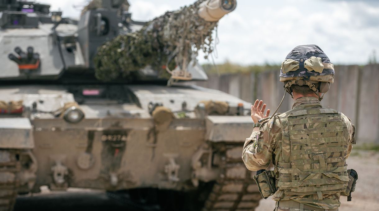 A soldier in camouflage uniform signals in front of a tank with a camouflaged barrel on a clear day.