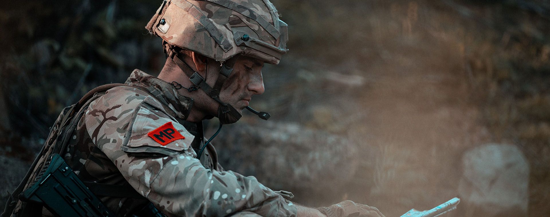 Camouflaged soldier wearing helmet and MP patch studies a map while seated, rifle slung across his back.