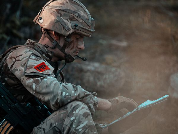 Camouflaged soldier wearing helmet and MP patch studies a map while seated, rifle slung across his back.