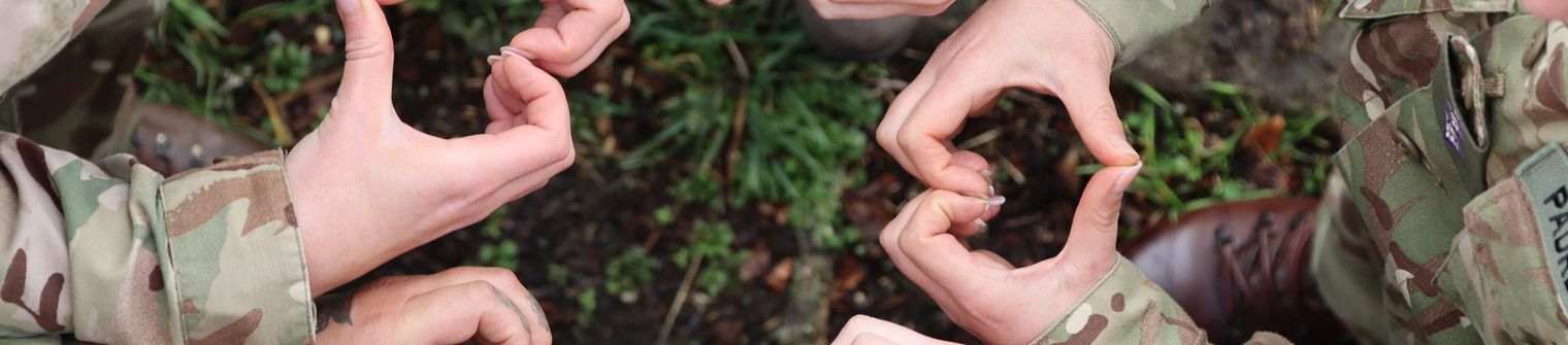 People in camouflage uniforms form heart shapes with their hands in a circle, symbolizing unity and friendship.