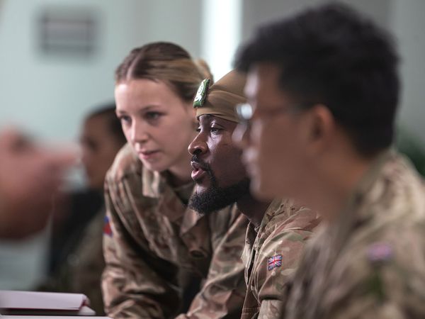 Three soldiers in camouflage uniforms, a woman leaning over, engaged in a focused discussion at a table.