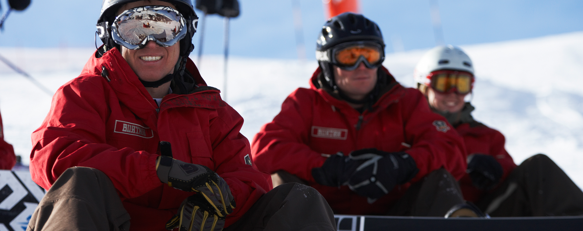 Three snowboarders in red jackets sitting on snow, smiling, wearing helmets and goggles, with snowboards in front of them and a snowy background.