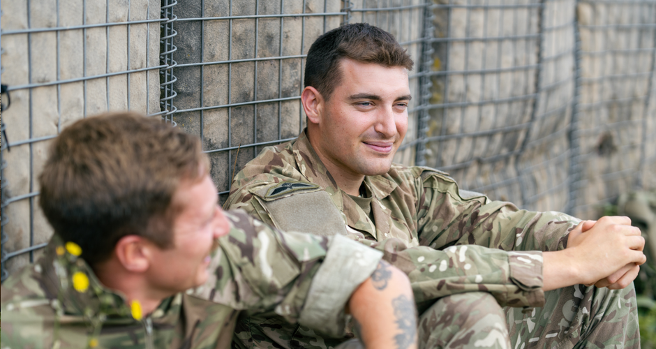 Two soldiers in camouflage uniforms sit and relax against a wire fence, smiling and engaged in conversation.