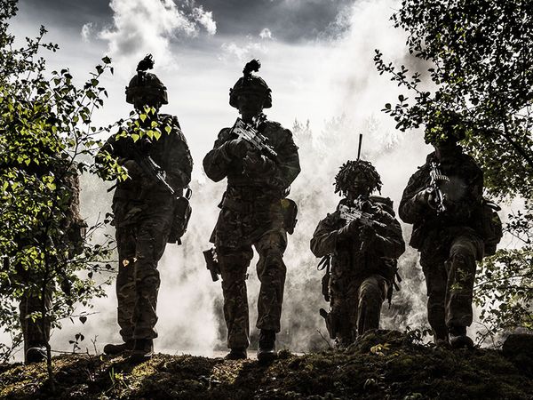 Silhouetted soldiers in uniform holding rifles, standing amid smoke and trees under a cloudy sky.
