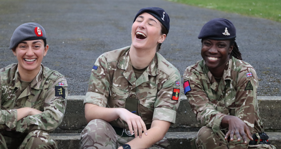 Three soldiers in camouflage uniforms sit on steps, smiling and laughing, wearing different colored berets.