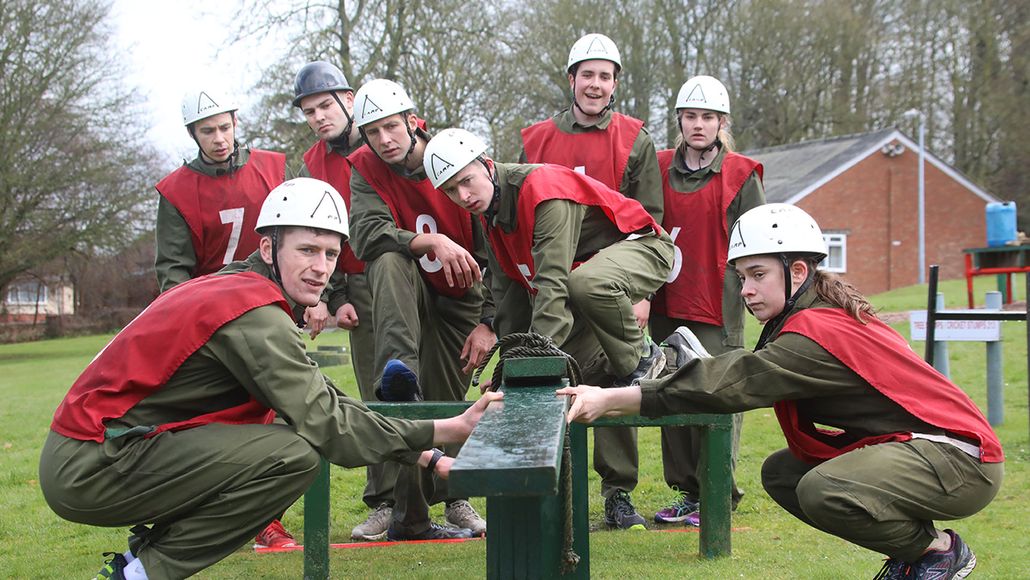 A group of people in red vests and white helmets, crouching around a green obstacle course outdoors, in a grassy area with trees in the background.
