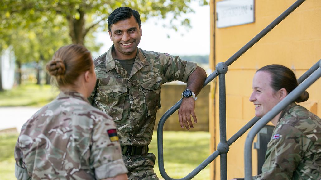 Three military personnel in camouflage uniforms converse outdoors near stairs, with trees and a yellow building in the background.