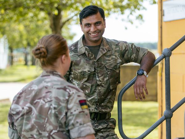 Three military personnel in camouflage uniforms converse outdoors near stairs, with trees and a yellow building in the background.