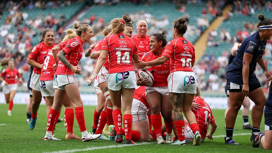 A women's rugby team in red uniforms celebrates a successful play on a grassy field with a cheering crowd in the background.