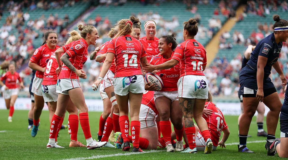 A women's rugby team in red uniforms celebrates a successful play on a grassy field with a cheering crowd in the background.