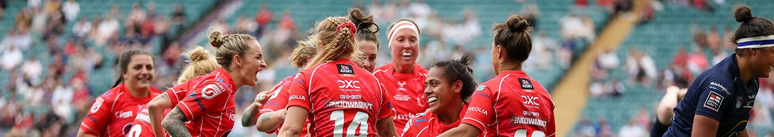 A women's rugby team in red uniforms celebrates a successful play on a grassy field with a cheering crowd in the background.