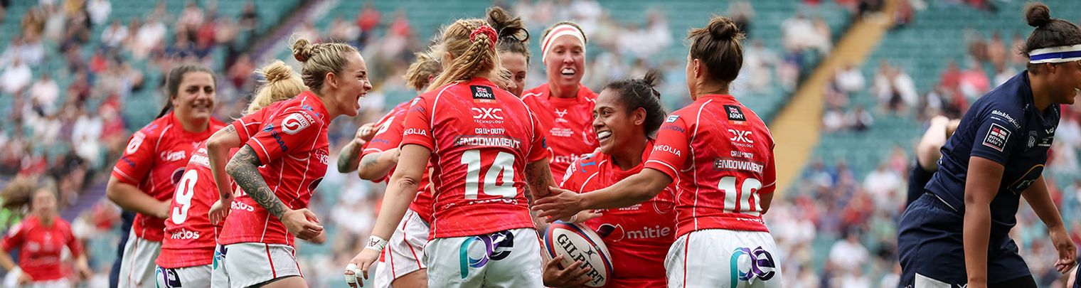 A women's rugby team in red uniforms celebrates a successful play on a grassy field with a cheering crowd in the background.
