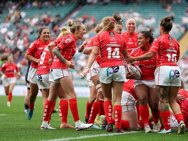 A women's rugby team in red uniforms celebrates a successful play on a grassy field with a cheering crowd in the background.
