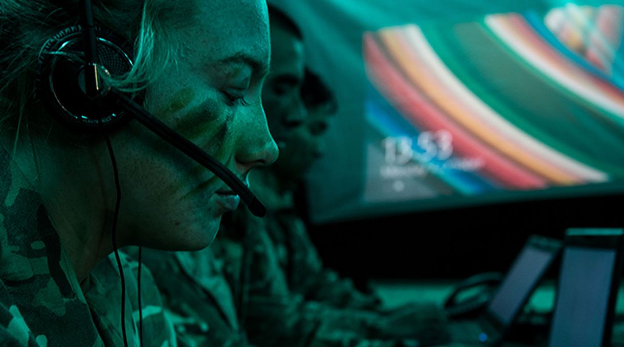 People in military uniform and face paint work at computers with headsets in a dimly lit, green-tinted room.