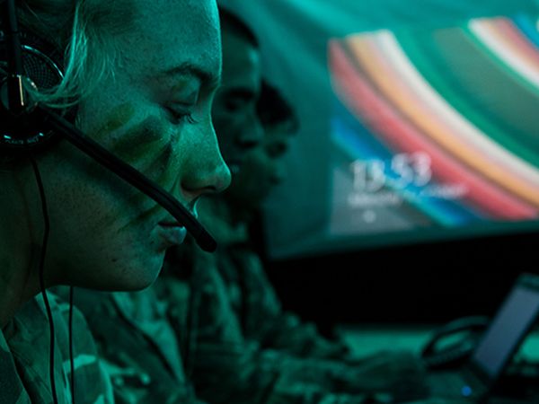 People in military uniform and face paint work at computers with headsets in a dimly lit, green-tinted room.