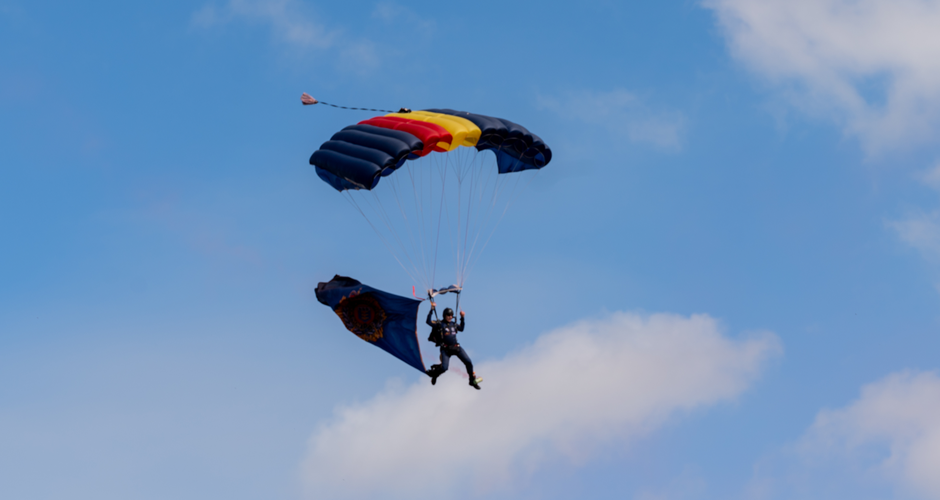 A parachutist descends through a blue sky with a colorful parachute and a large flag, displaying a circular emblem.