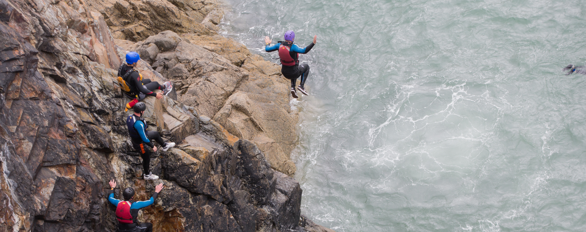 People in helmets and wetsuits climbing rocky cliffside near choppy water, with one person jumping into the water.