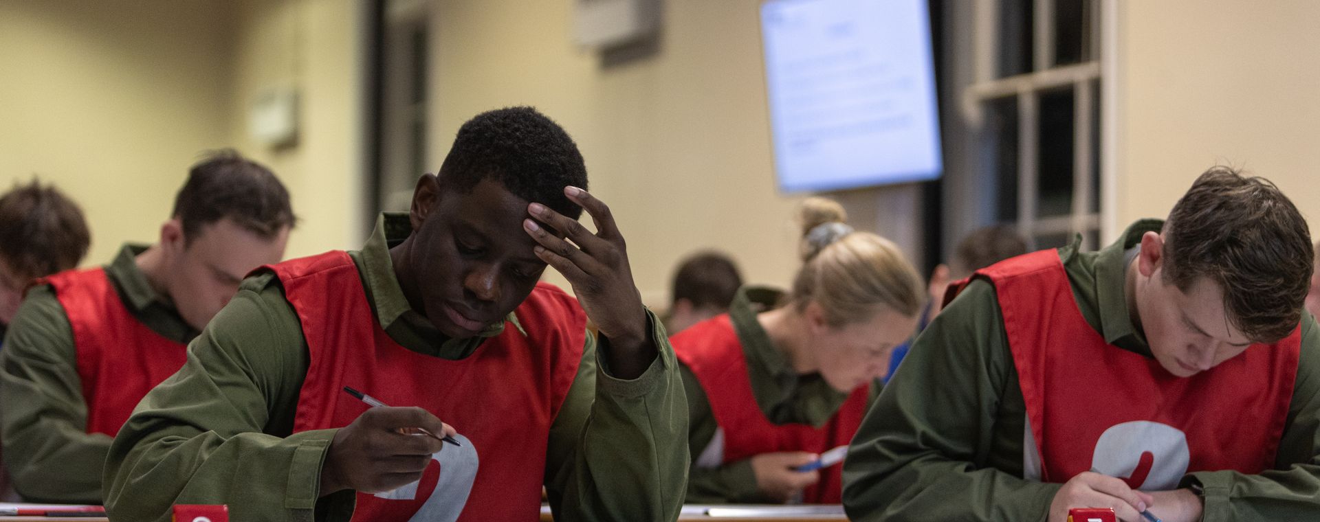 People wearing red numbered vests concentrate on a written exam in a classroom setting.