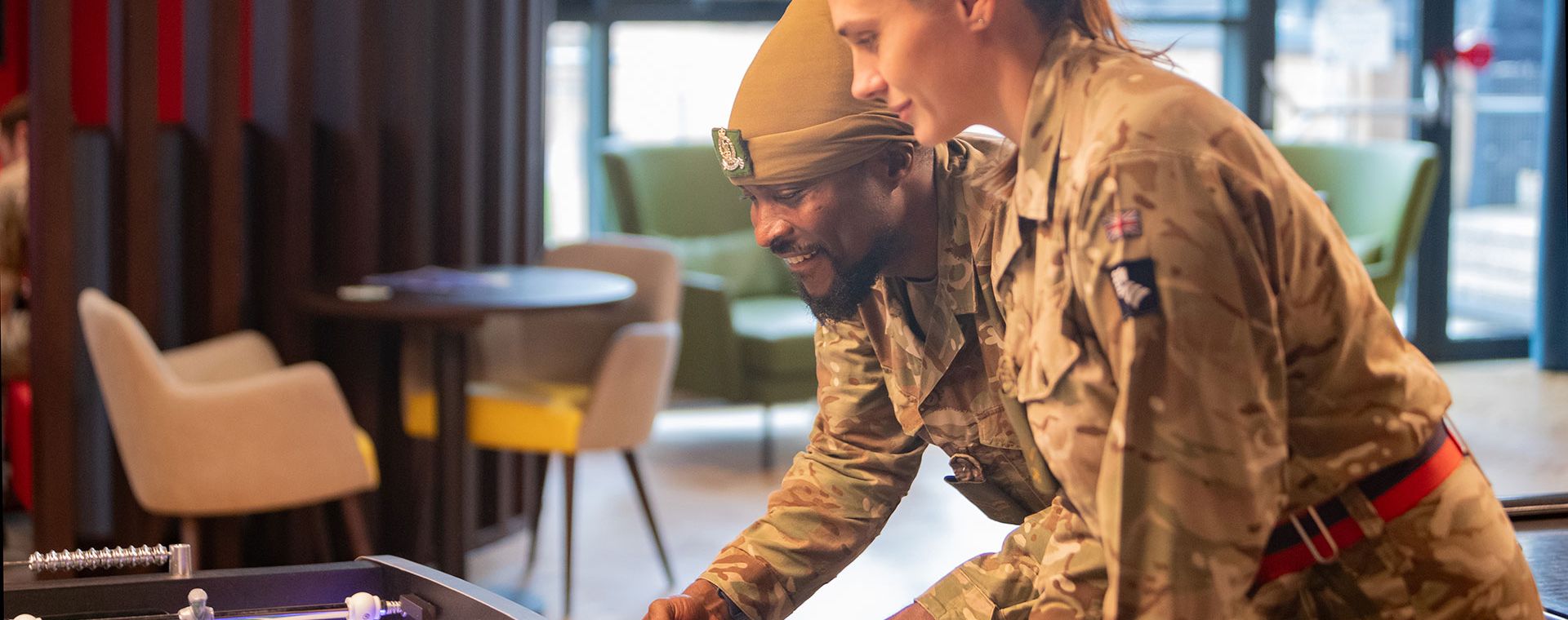 Two soldiers in uniform play foosball together, smiling and focused in a recreational room with chairs and a table in the background.